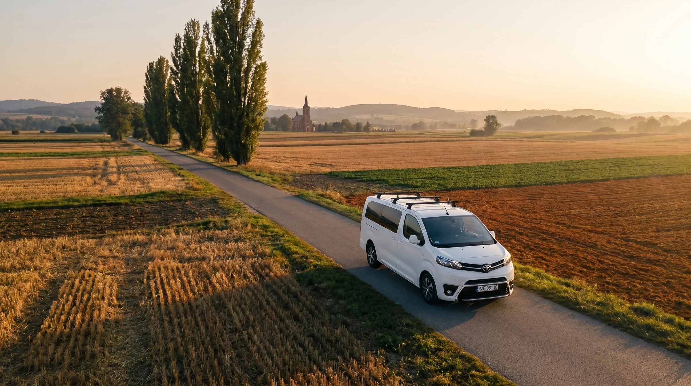 Biały bus Toyota Proace Verso jadący wiejską drogą o złotej godzinie, w tle polskie pola i kapliczka